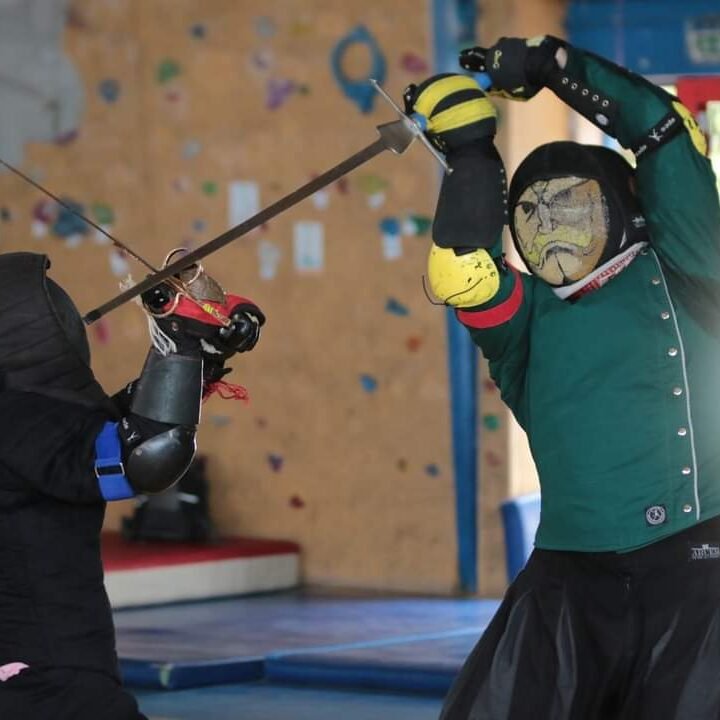 deux amheurs en train de combattre. L'un habillé de vert, de jaune et de noir, avec une masque figurant le portrait de Alex Louis Armstrong porte un coup plongeant depuis la posta della fenestra